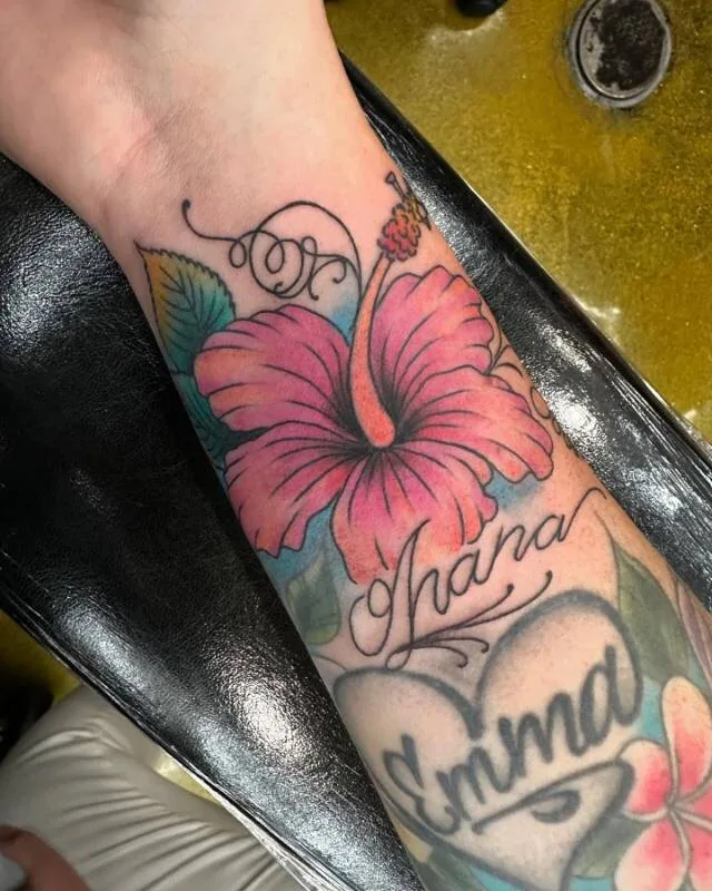 Illustrative color memorial tattoo with hot-pink hibiscus, flowing 'Ohana' script and an 'Emma' heart-locket on the forearm and wrist, by Julian (xwildxlovex) at Ohana Tattoo, Waikiki, Hawaii.