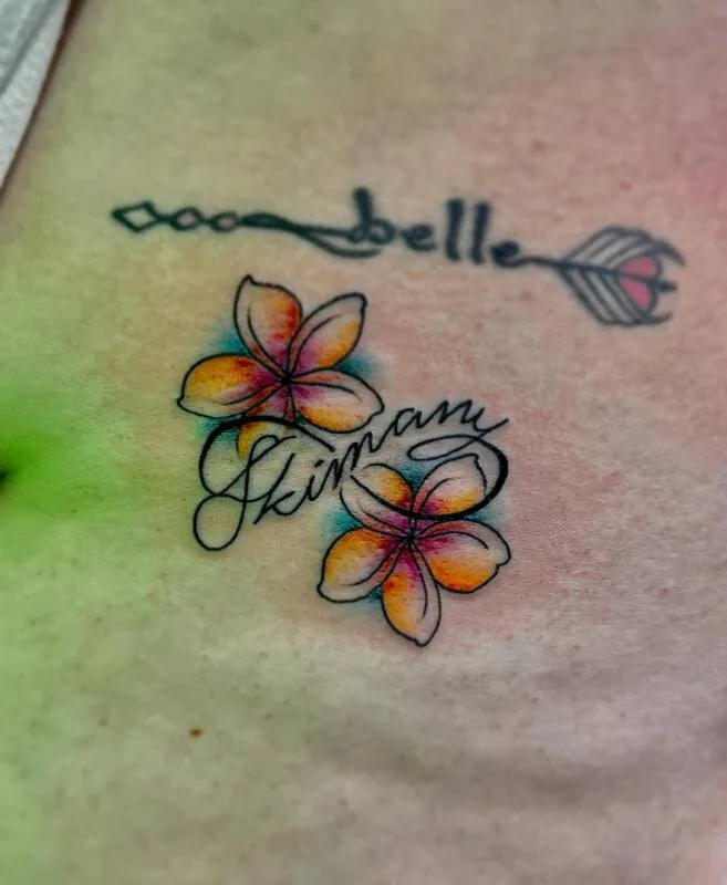 Illustrative color tattoo of two plumeria flowers with flowing 'Skimani belle' script on the ribs, by Julian (xwildxlovex) at Ohana Tattoo, Waikiki, Hawaii.